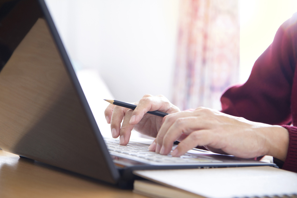 Hands of a man in red sweater holding a pencil while typing on laptop computer notebook keyboard in the office. Closeup with left and top copy space and easy to be cropped as a banner or strip