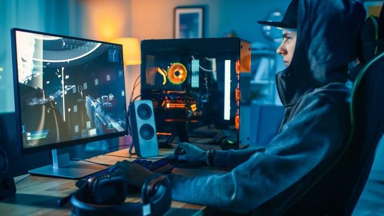 Professional Gamer and Streamer Playing First-Person Shooter Online Video Game on His Cool Personal Computer. Young Man is Wearing a Cap and Hood. Room and PC have Colorful Neon Led Lights. Cozy Evening at Home.